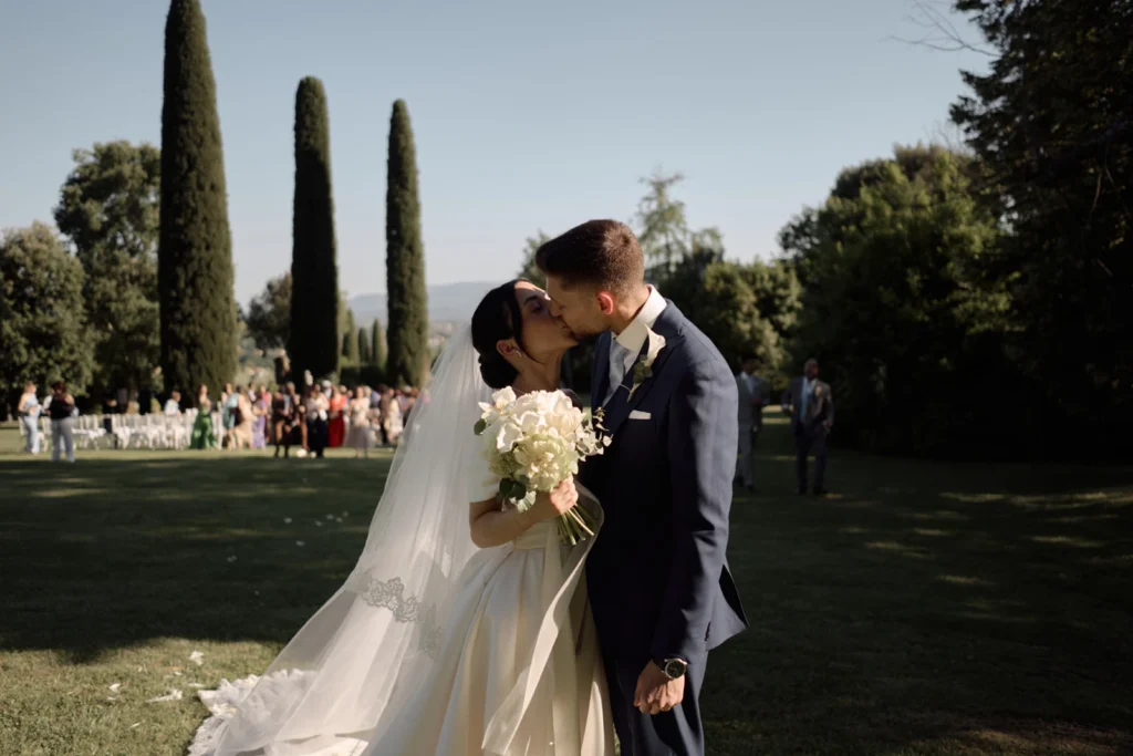 Sposi si baciano sul prato dopo la cerimonia di matrimonio in Toscana, bouquet di ortensie bianche e cipressi sullo sfondo