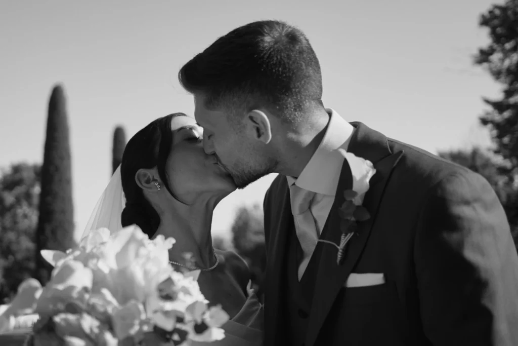 Ritratto in bianco e nero degli sposi che si baciano dopo la cerimonia di matrimonio in Toscana, con bouquet bianco e cipressi