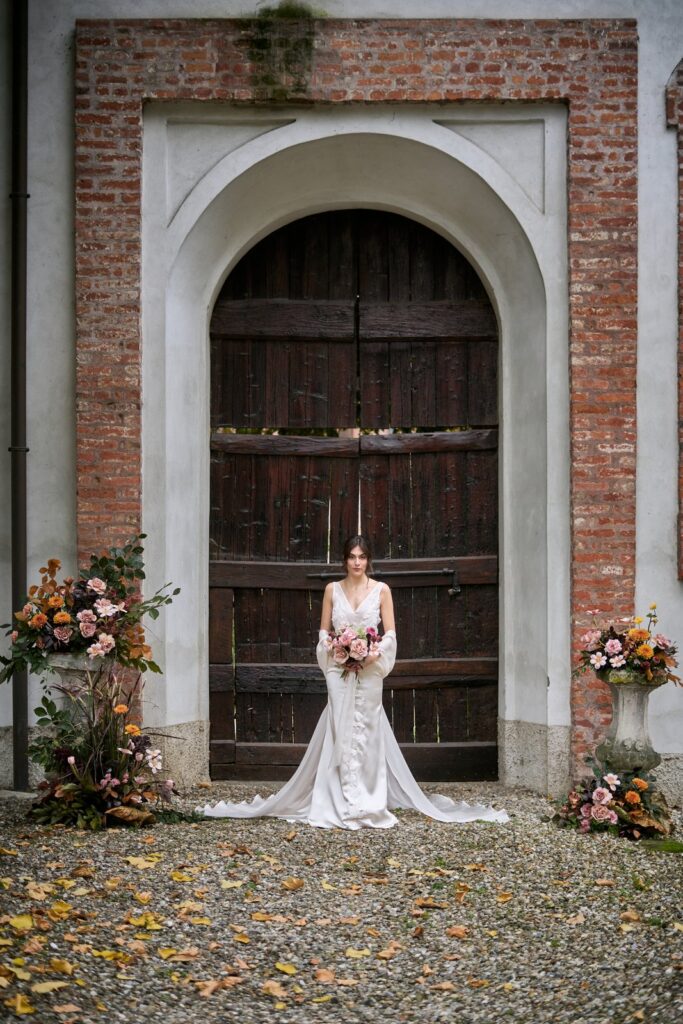 Sposa con abito in raso e strascico e bouquet autunnale davanti a un portone in legno